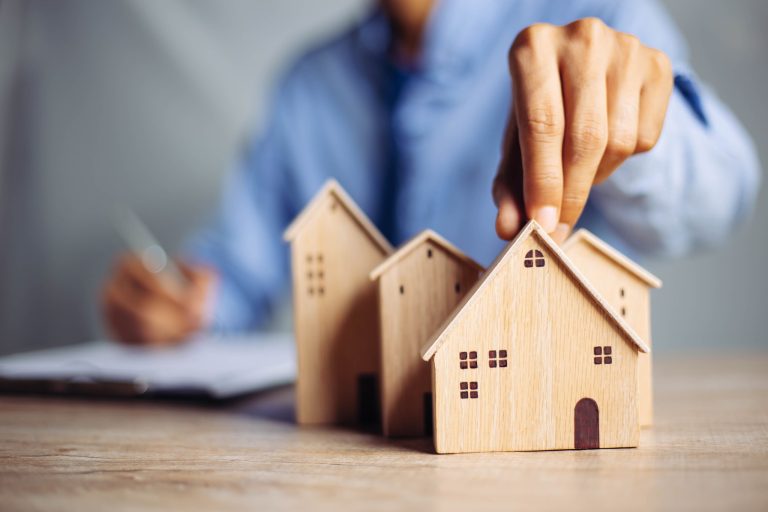 A hand places a small wooden house model beside paperwork, illustrating property valuation